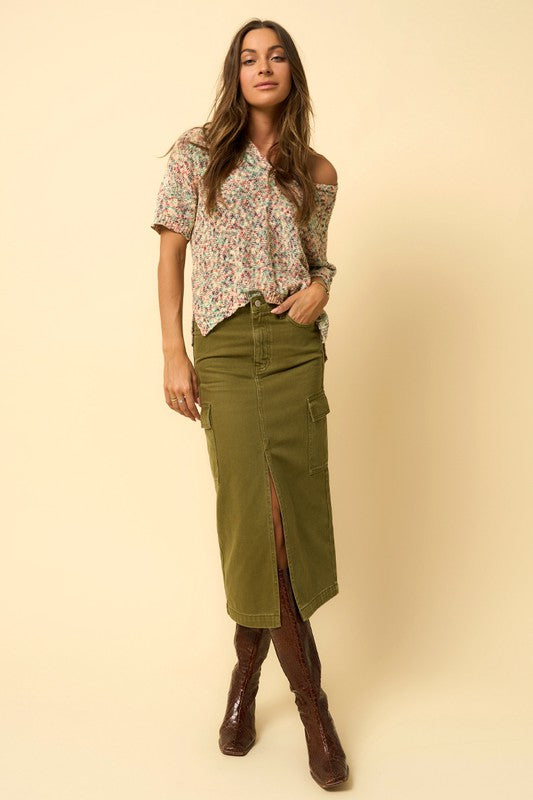 Model standing with hands in pockets wearing olive midi denim skirt and floral blouse.
