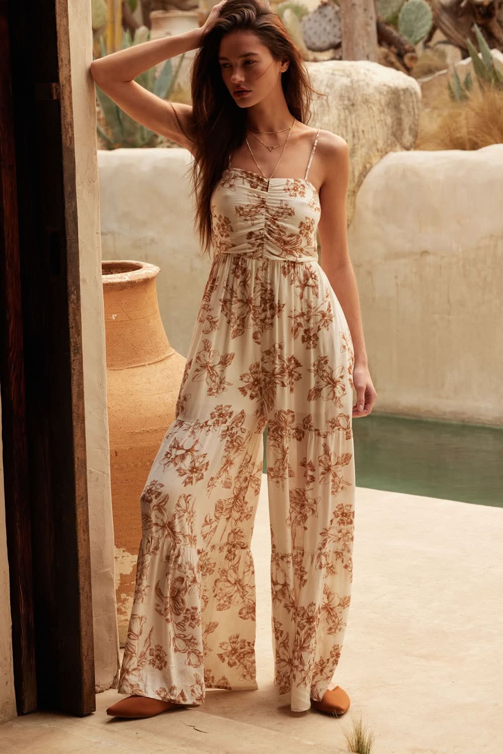 AVAH-Model wearing taupe floral print wide-leg jumpsuit with ruched sweetheart bodice, adjustable straps, and flowing crepe fabric by a poolside doorway.
