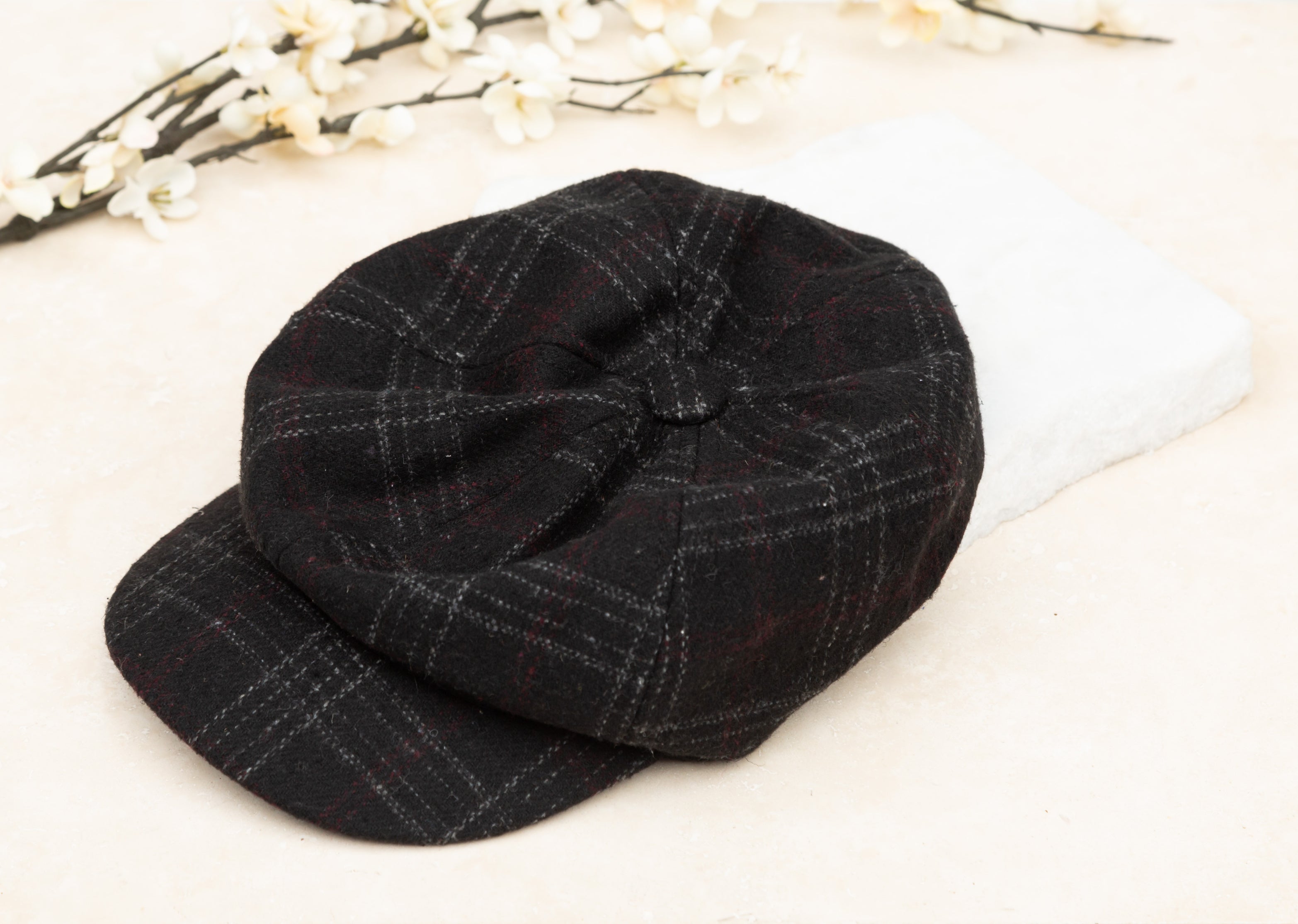 A black tweed baker boy cap with a structured brim, flat lay on white background
