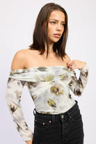 Model posing in AVAH off-the-shoulder ruched mesh bodysuit with long sleeves in an olive floral print.