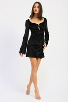 Walking pose in black mini dress with sweetheart neckline and centered gold clasp.
