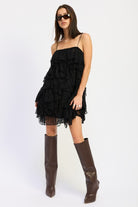 Model posing in black tiered lace mini dress with layered ruffles and boots.
