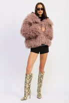 Model wearing the mauve textured coat pulled close at the front with high boots and black shorts.