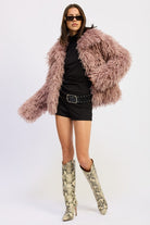 Model showcasing the coat open over black shorts and a studded belt with knee-high boots.