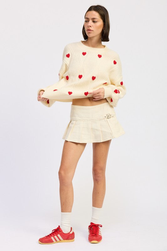 Ivory knit sweater with red heart details and textured pattern, styled with cream pleated mini skirt and red shoes