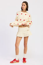 Ivory knit sweater with red heart details and textured pattern, styled with cream pleated mini skirt and red shoes