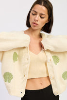Close-up of ivory knit cardigan showing green embroidered shells and button detail.
