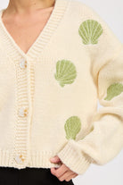Close-up of ivory knit cardigan showing green embroidered shells and button detail.
