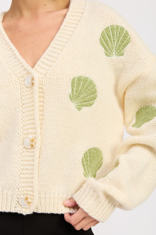 Close-up of ivory knit cardigan showing green embroidered shells and button detail.
