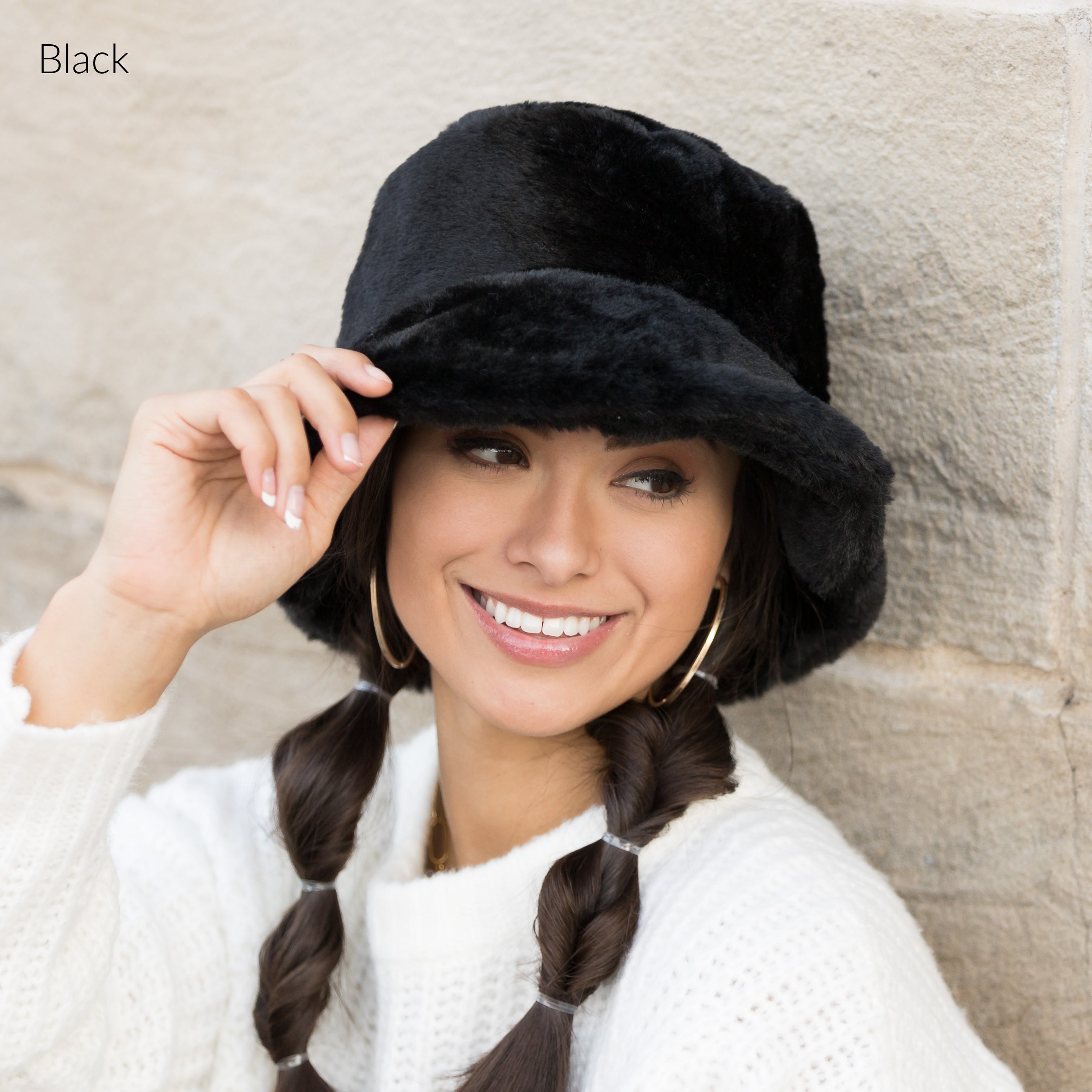 Woman wearing a black plush faux fur bucket hat, holding the brim, side view against a stone wall