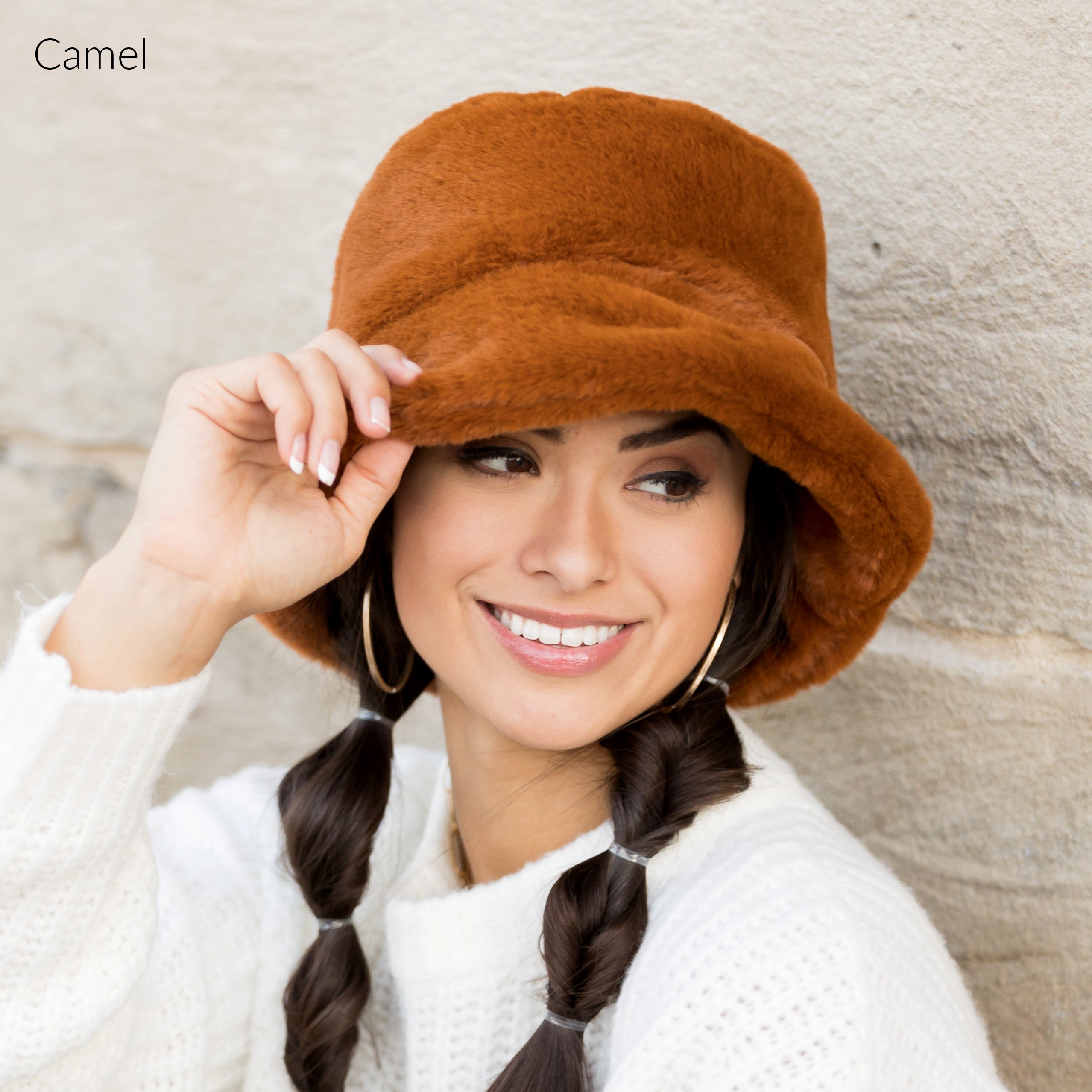 Woman wearing a camel plush faux fur bucket hat, holding the brim, side view against a stone wall