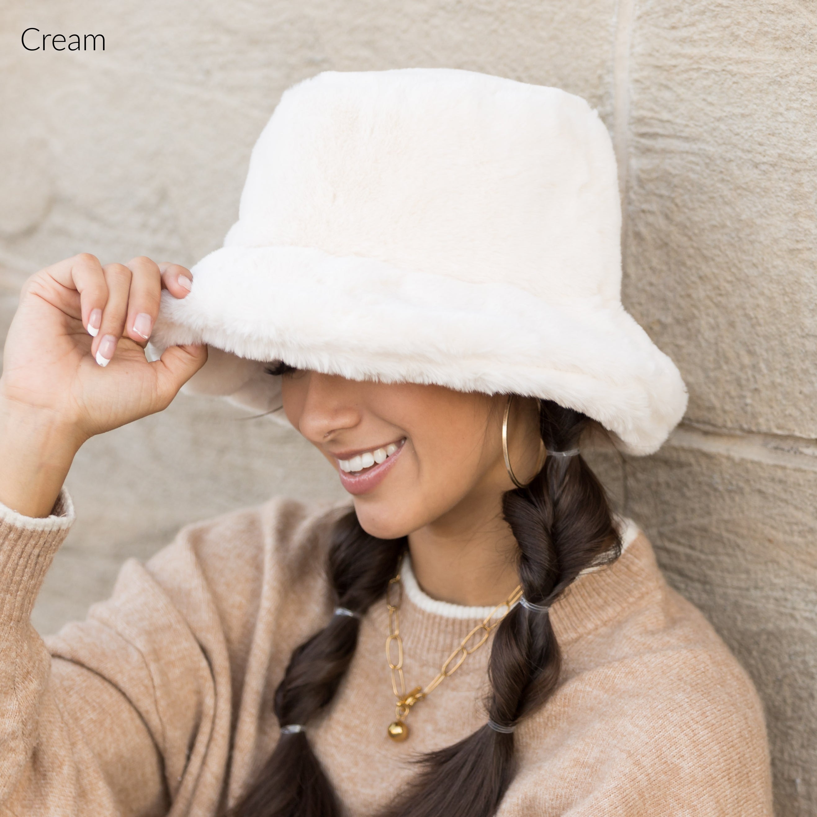 Woman wearing a white plush faux fur bucket hat, holding the brim, side view against a stone wall