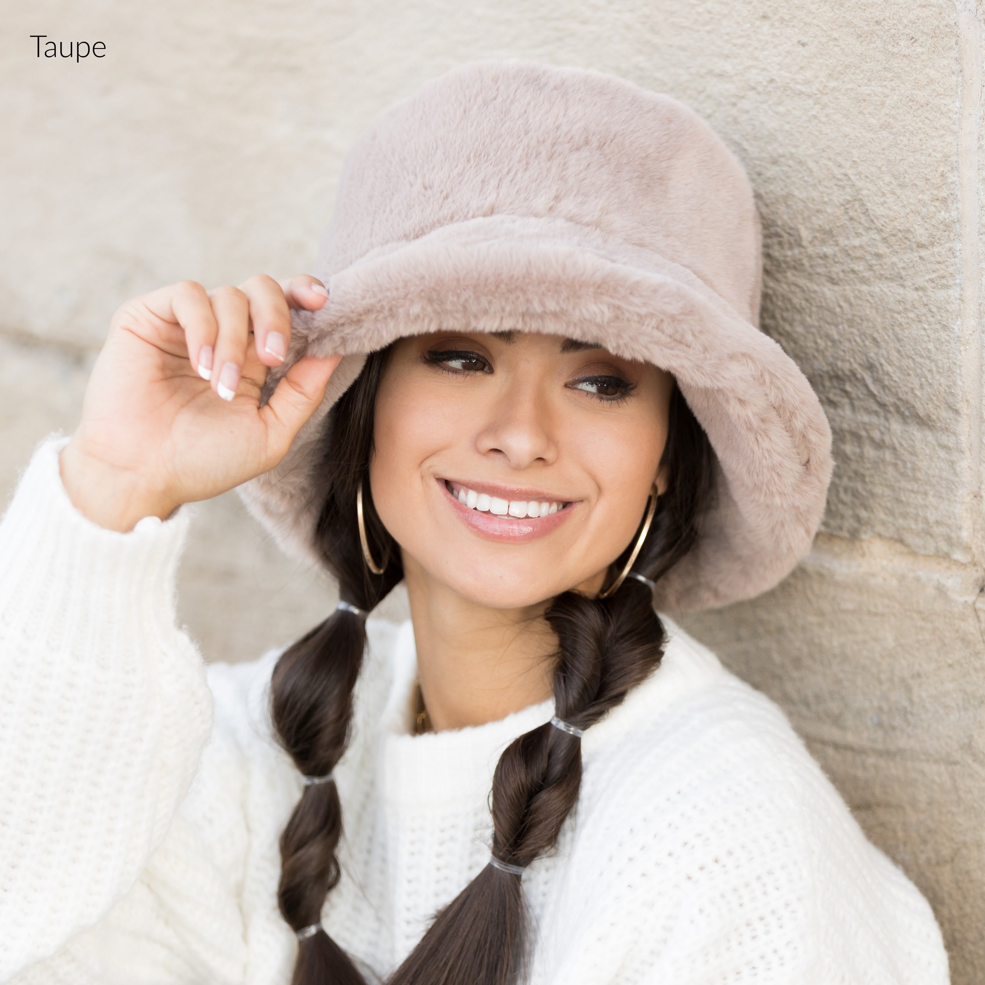 Woman wearing a taupe plush faux fur bucket hat, holding the brim, side view against a stone wall