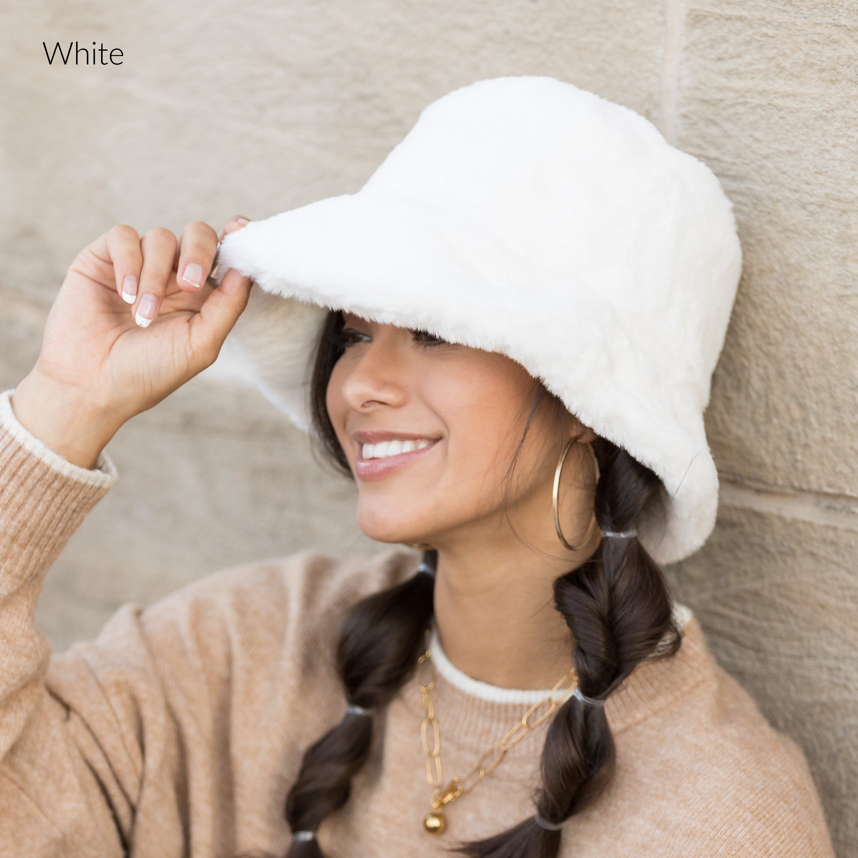 Woman wearing a white plush faux fur bucket hat, holding the brim, side view against a stone wall