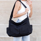 Person carrying a black shoulder messenger bag with two outer flap pockets and two open end pockets against a beige wall