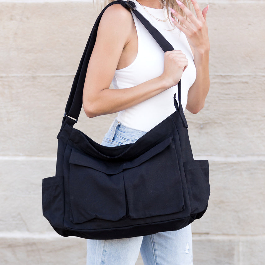 Person carrying a black shoulder messenger bag with two outer flap pockets and two open end pockets against a beige wall