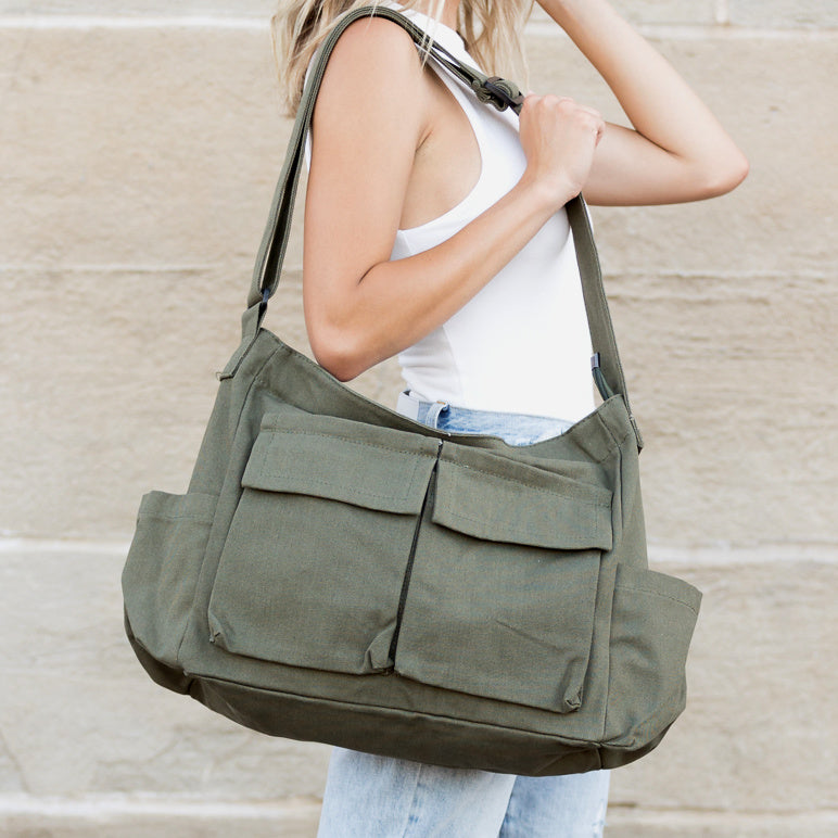 Person holding an olive green shoulder messenger bag with two outer pockets and two open end pockets against a beige wall