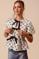 Front view of woman wearing a textured cream peplum top with black ribbon bow ties and puff sleeves | AVAH Collections