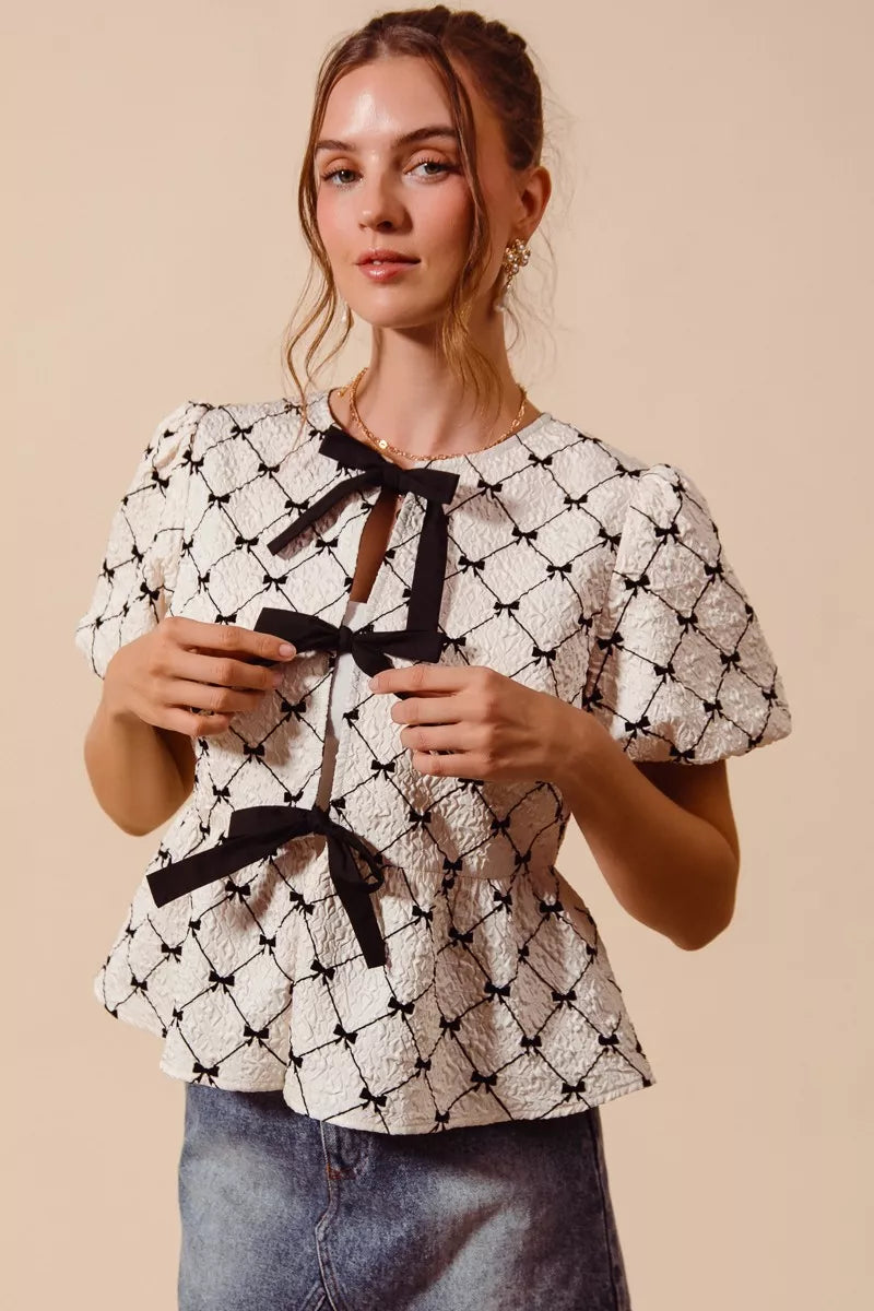 Front view of woman wearing a textured cream peplum top with black ribbon bow ties and puff sleeves | AVAH Collections