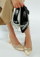 Model wearing beige satin block heels with crystal embellishment, holding black and white pairs

