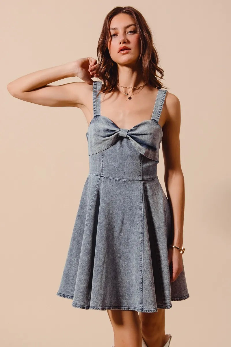 Model wearing a light wash denim mini dress with bow front neckline and fit and flare silhouette
