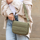 Person wearing a beige coat and holding an olive crossbody quilted puff foldover bag against a neutral background
