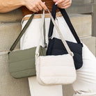 Person holding three quilted handbags: a green, a black, and a beige one, against a stone wall background.