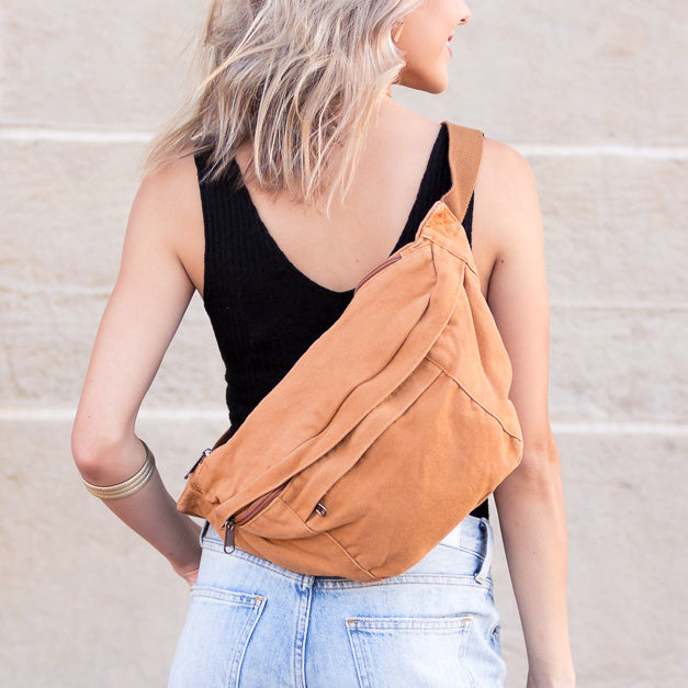 A woman wearing a black ribbed tank top and light blue jeans, shown from the back and side, wearing a camel canvas oversized crossbody sling bag against a light beige brick wall.