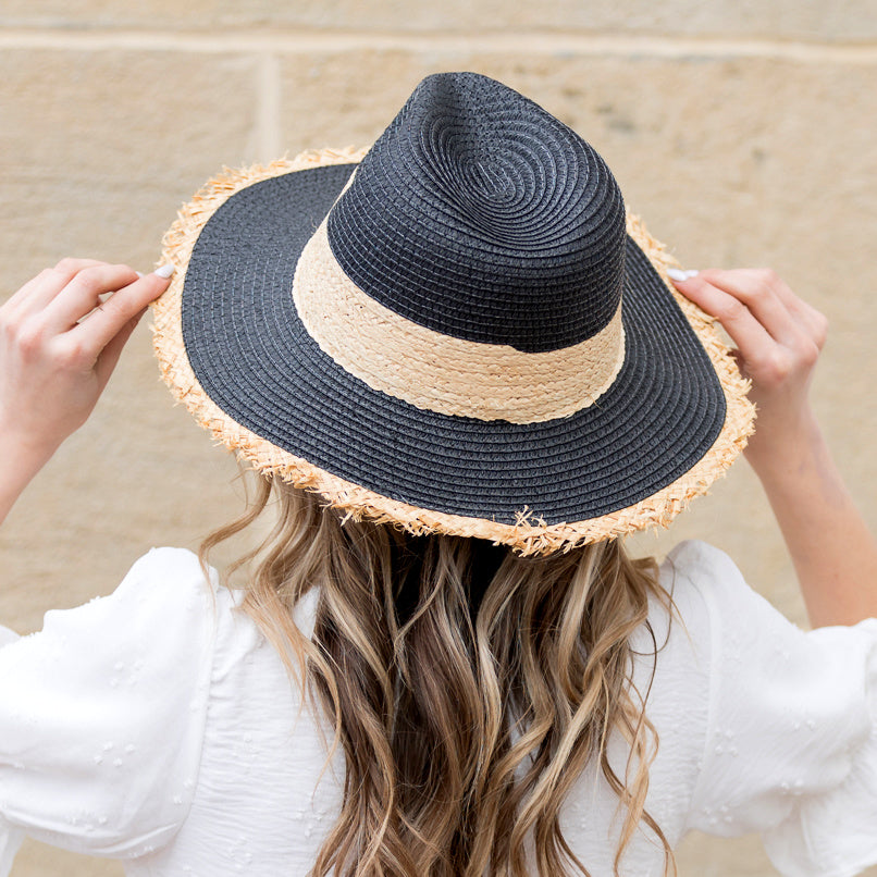 Back view of woman wearing a black straw sun hat with natural rattan band and frayed fringe brim
