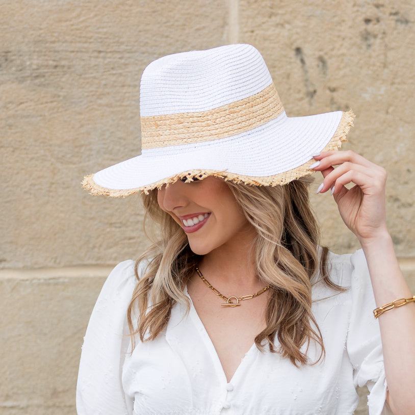 Side view of woman wearing a white straw sun hat with natural rattan band and touching the frayed fringe brim

