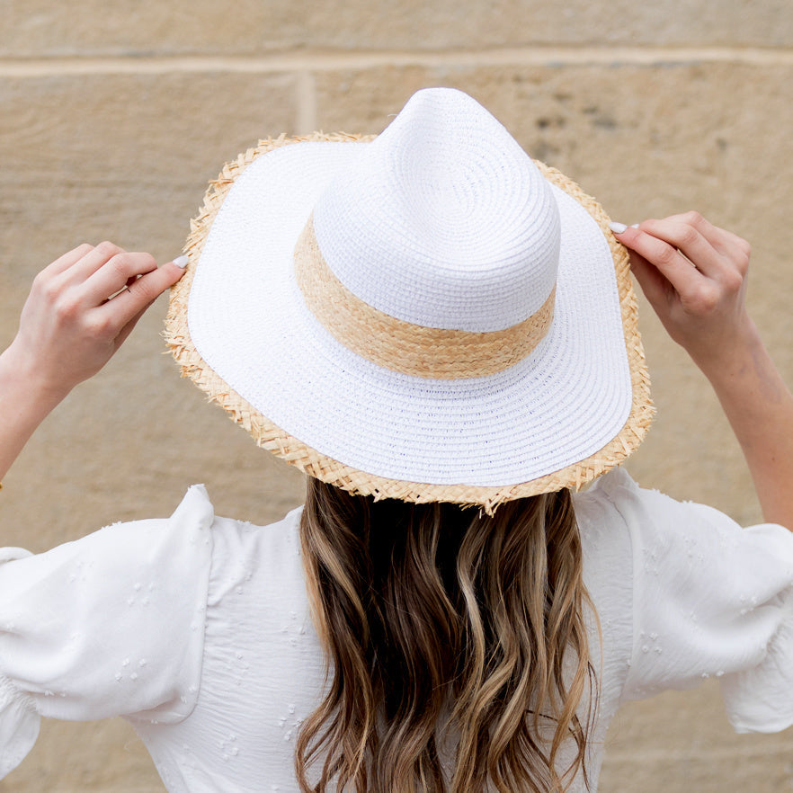 Back view of woman wearing a white straw sun hat with natural rattan band and holding the frayed fringe brim