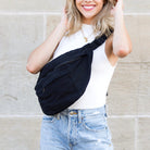 Woman wearing a black canvas oversized crossbody sling bag over her shoulder against a beige wall.