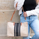 Person holding a beige tote bag with black vertical center stripe with brown straps against a stone wall.
