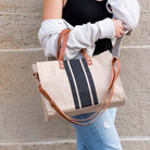 A woman wearing a white knit sweater and light blue jeans carries a canvas tote bag with a beige body, black and white vertical stripes, and brown leather handles and shoulder strap against a stone wall.