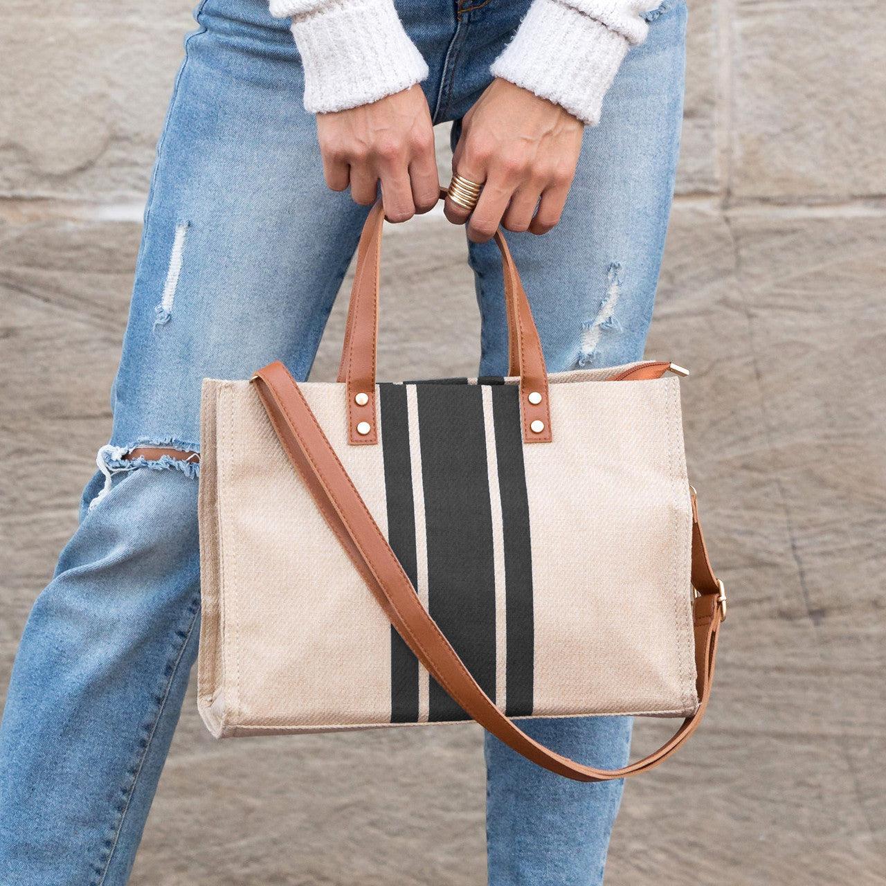 Person holding a striped tote bag with brown leather straps against a stone wall background