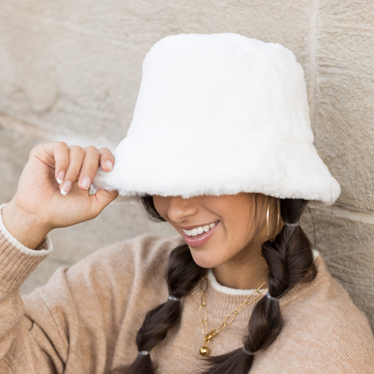 Woman wearing a white plush faux fur bucket hat, holding the brim, side view against a stone wall