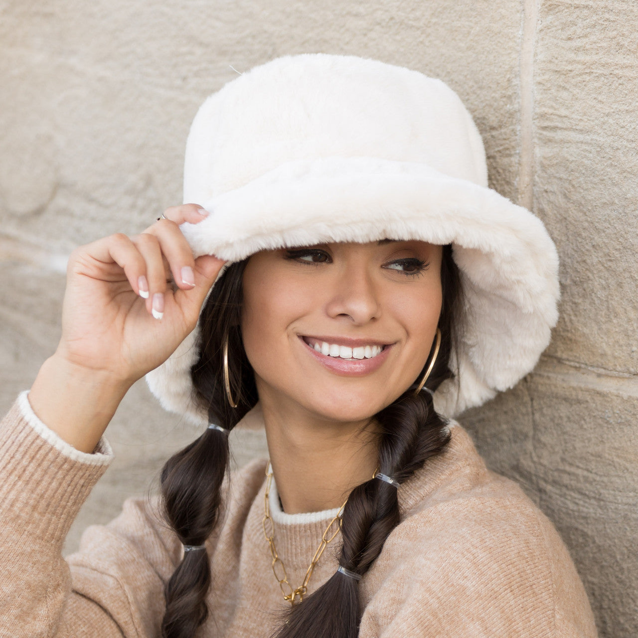 Woman wearing a cream plush faux fur bucket hat, holding the brim, side view against a stone wall