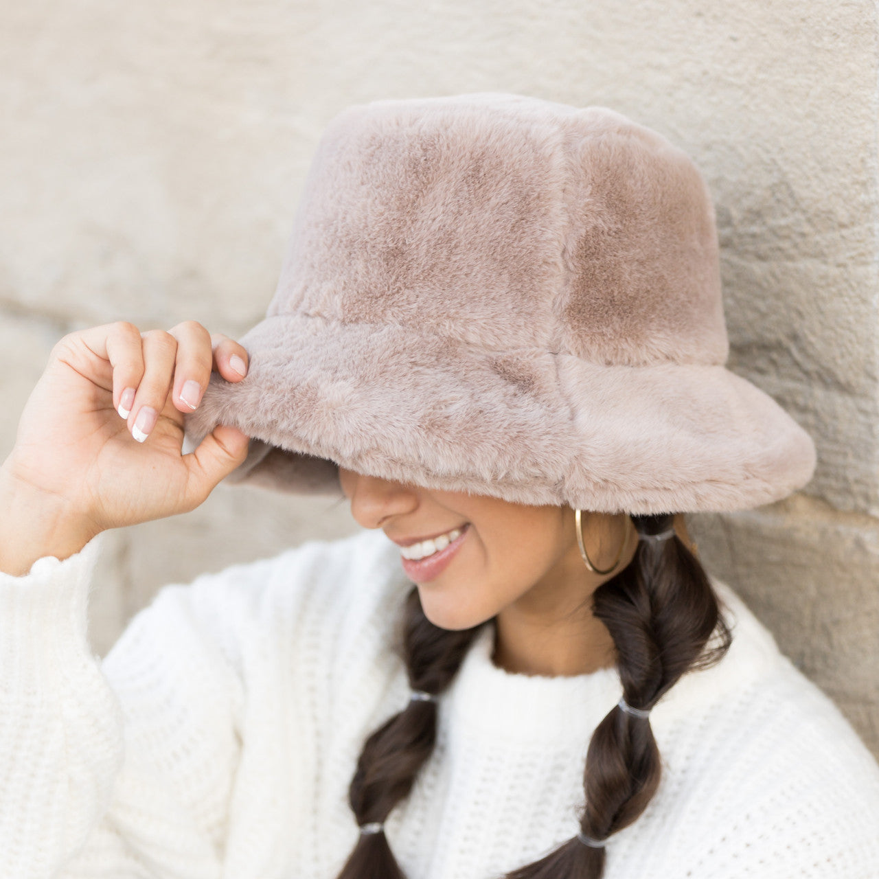 Woman wearing a taupe plush faux fur bucket hat, holding the brim, side view against a stone wall