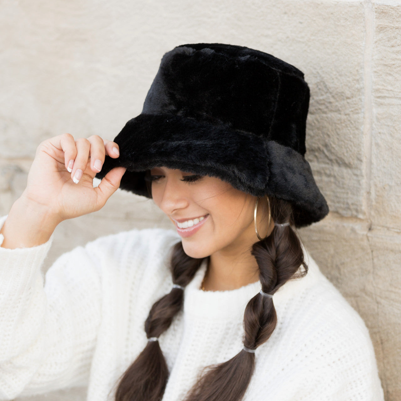 Woman wearing a black plush faux fur bucket hat, holding the brim, side view against a stone wall