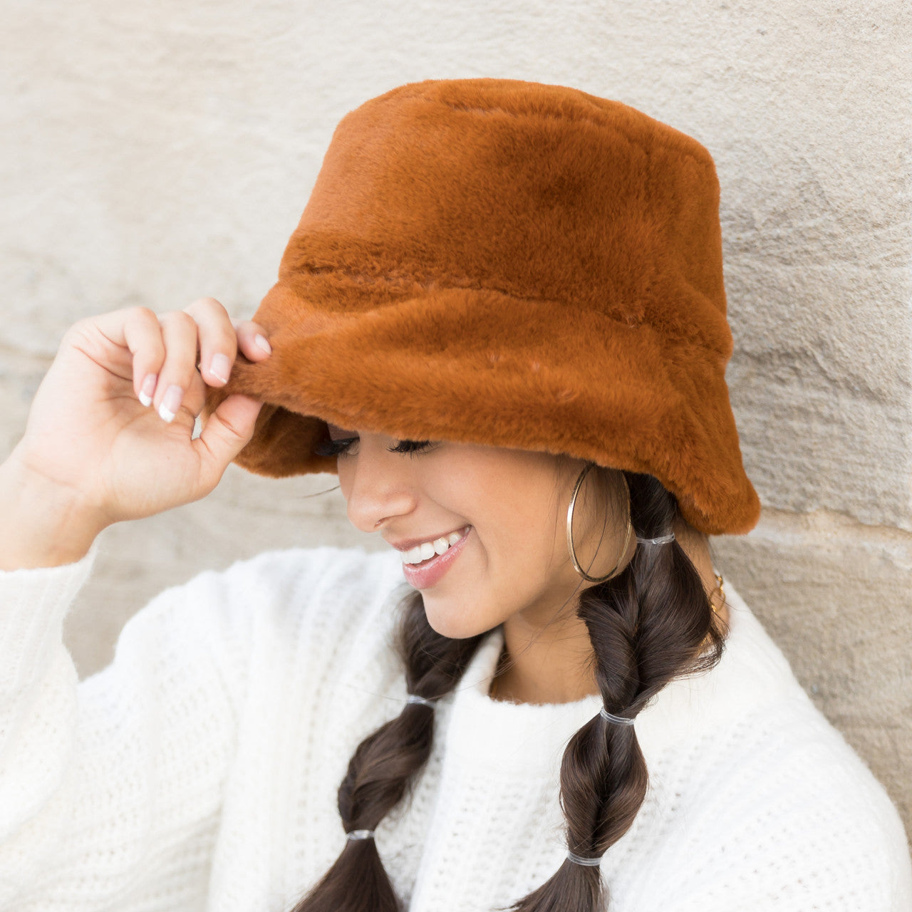 Woman wearing a camel plush faux fur bucket hat, holding the brim, side view against a stone wall