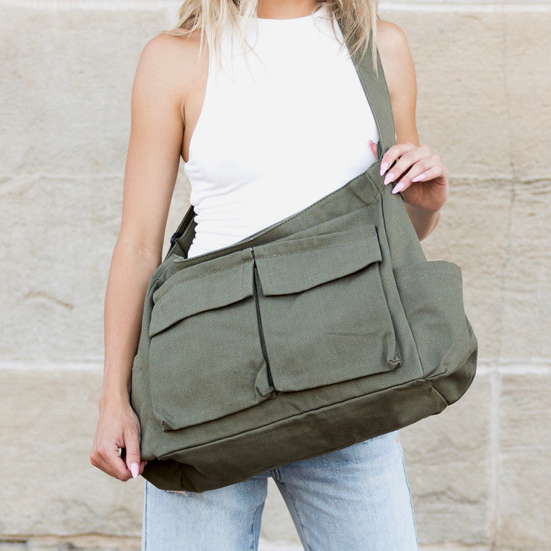 Person holding an olive green shoulder canvas messenger bag against a stone wall background