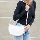 Person wearing a black long-sleeve top and light blue jeans, holding a bone handbag against a beige wall.
