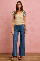 Woman wearing a beige crochet flutter sleeve top paired with wide leg denim jeans and platform sandals.
