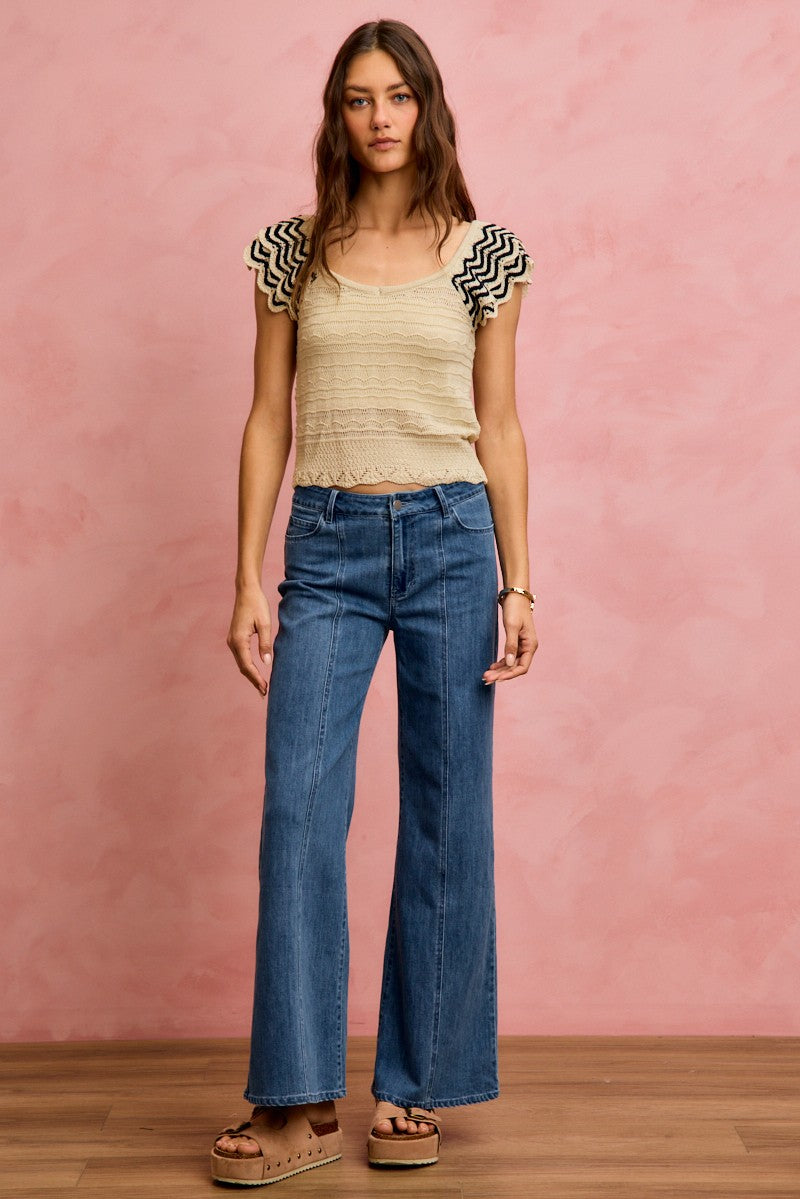Woman wearing a beige crochet flutter sleeve top paired with wide leg denim jeans and platform sandals.

