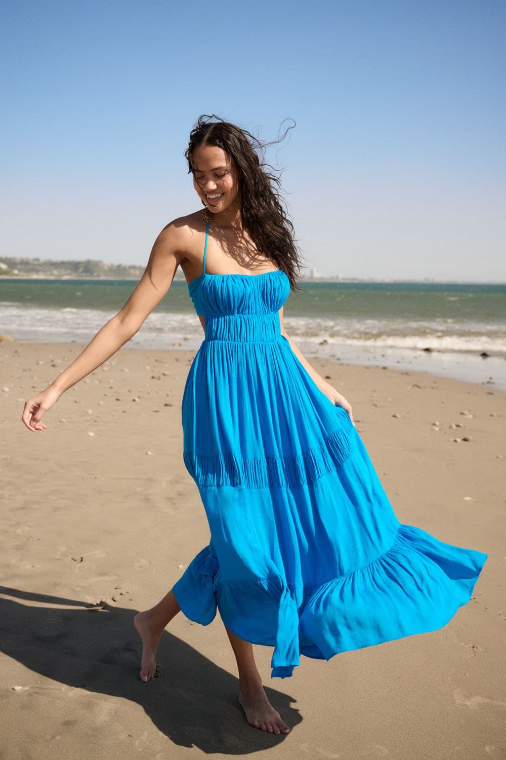 Model walking barefoot on a beach in a bright blue halter neck maxi dress with gathered bodice and flared tiered skirt.-AVAH