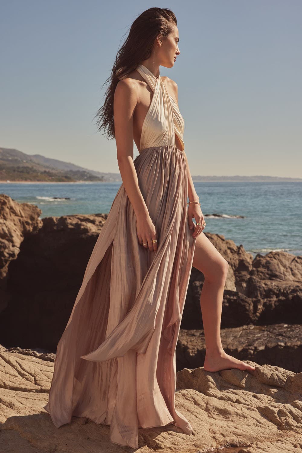 AVAH-Model standing barefoot on rocky coastline wearing cream and taupe halter neck satin maxi dress with crisscross bodice and high slit