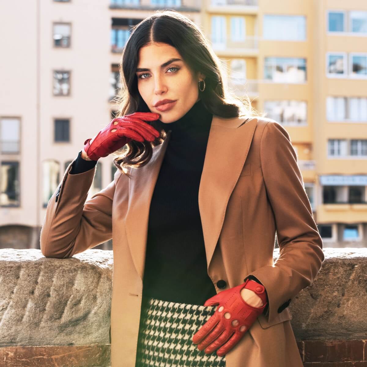 Woman in a tan coat and red leather drivig gloves standing in an urban setting