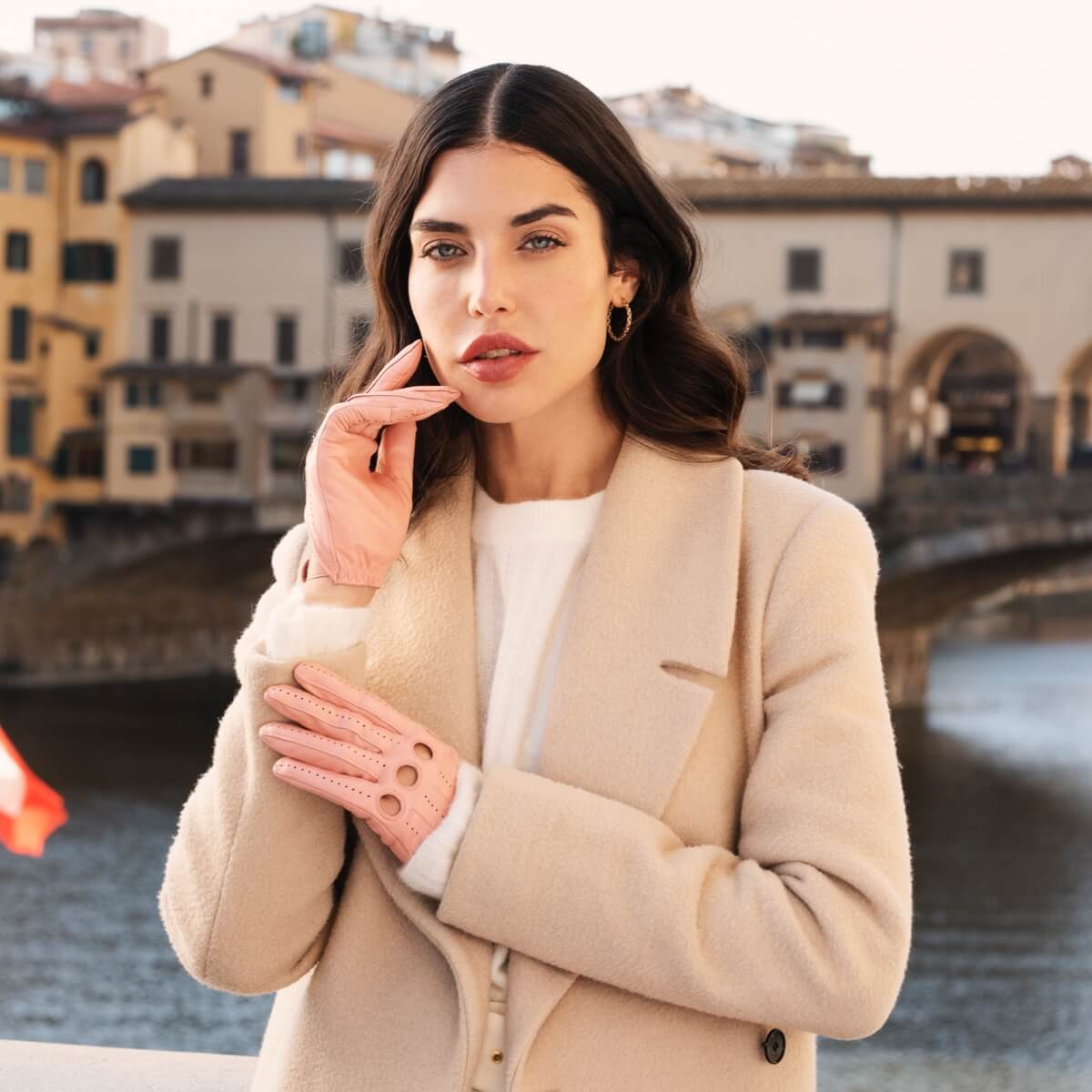 Woman in a beige coat with pink leather driving gloves standing by a river with buildings in the background