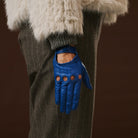 Blue leather glove worn by a person with a fur-lined hood and brown pants on a dark background