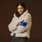 Woman wearing a beige fur coat and blue gloves against a brown background
